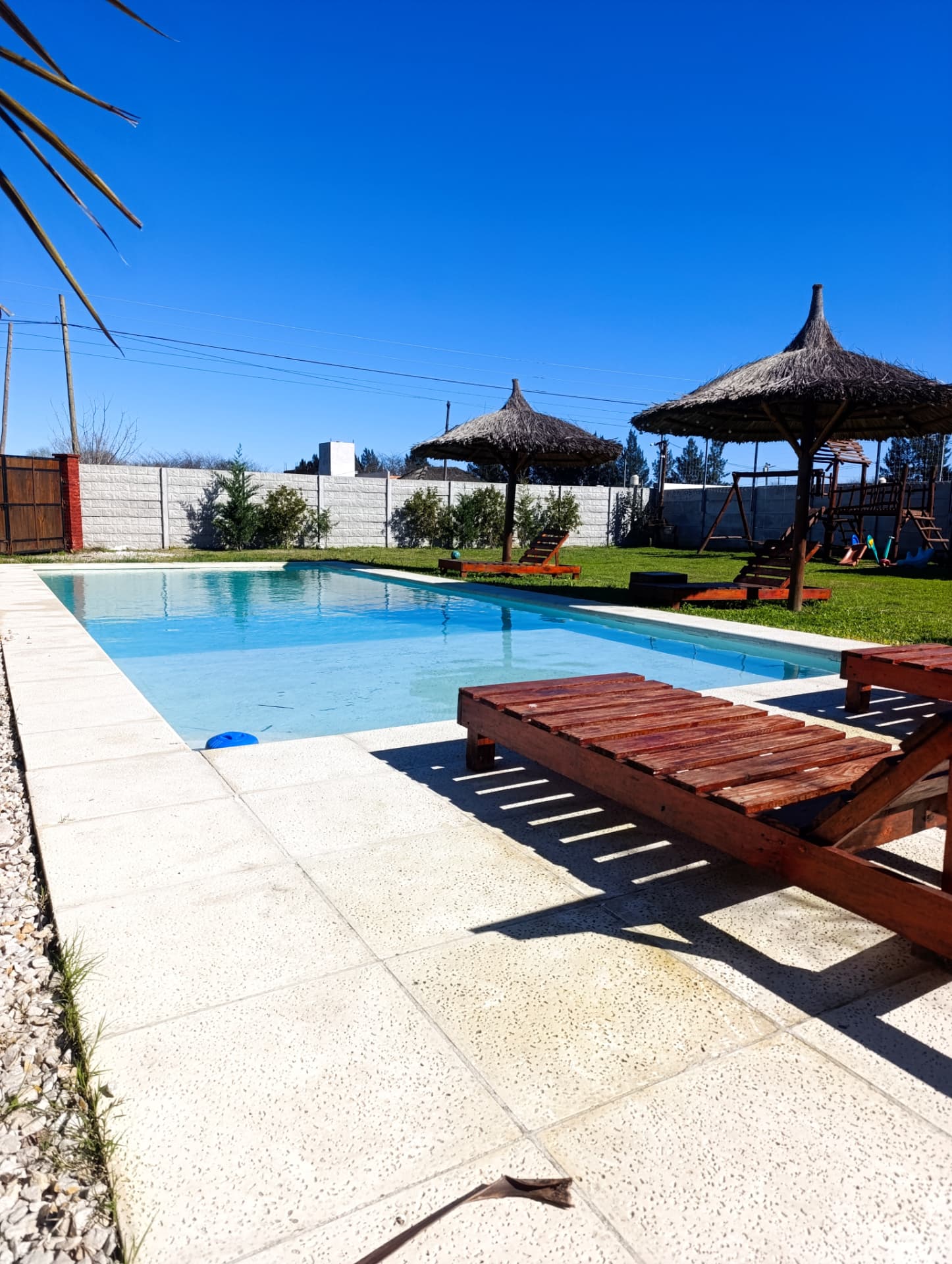 Piscina exterior con reposeras de madera y sombrillas de paja bajo cielo azul